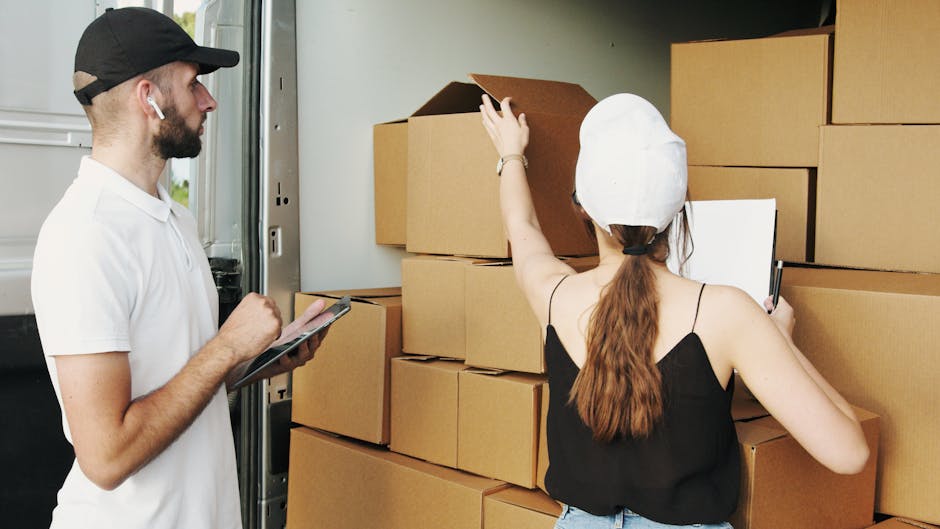 Two movers from Barnes Removals are engaged in a home relocation process involving the loading of cardboard boxes into the back of a moving van. The male mover, dressed in a white shirt, black cap, an