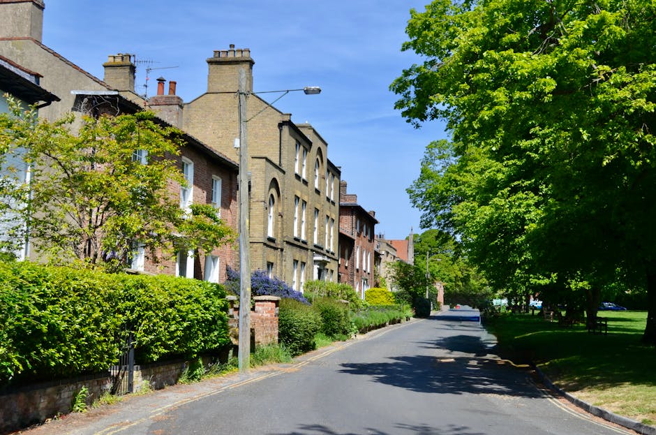 A residential street scene showing a row of Victorian-style brick houses with tall chimneys, large windows, and decorative facades on the left side of the image. The houses are partially obscured by lush green trees and well-maintained hedges, with some flowering plants visible near the houses. A narrow asphalt road curves gently to the right, lined with green grass and shaded by mature trees on the right side, creating a peaceful and leafy environment. A grey streetlamp is mounted on a tall pole near the center of the scene, and the sky above is clear and blue. This setting illustrates the typical environment in a suburban area suitable for house removals and home relocation services by Barnes Removals, as they assist with furniture transport and packing during moving logistics companies' operations.
