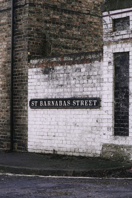 A street sign reading 'St Barnabas Street' affixed to a white brick wall on a corner of a building, with surrounding weathered brickwork in dark red and black hues. The sign is black with white lettering, positioned near the street level, with a sidewalk in front. The building's exterior shows signs of age and wear, consistent with an urban environment suitable for house removals and furniture transport. The area appears clean with minimal street debris visible, and natural daylight illuminates the scene. This setting may represent part of a residential or commercial area in Barnes, relevant to local house removals and moving services provided by Barnes Removals.
