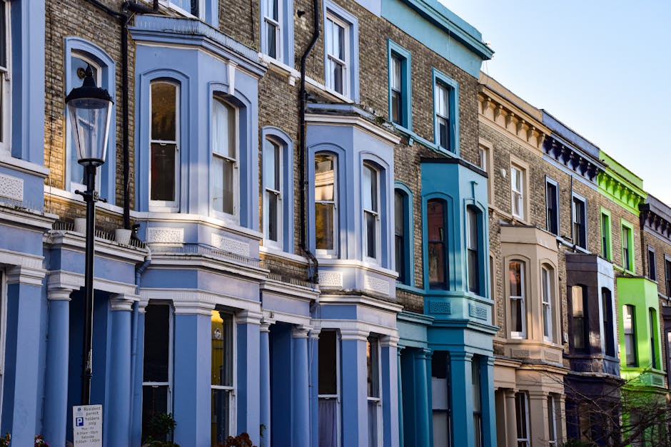 A residential street lined with large, mature trees with dense, green foliage extending over the pavement. The street features white, ornate Victorian-style terraced houses with decorative facades and